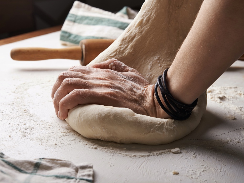 A hand kneading some dough on a floured surface, with a rolling pin in the background.