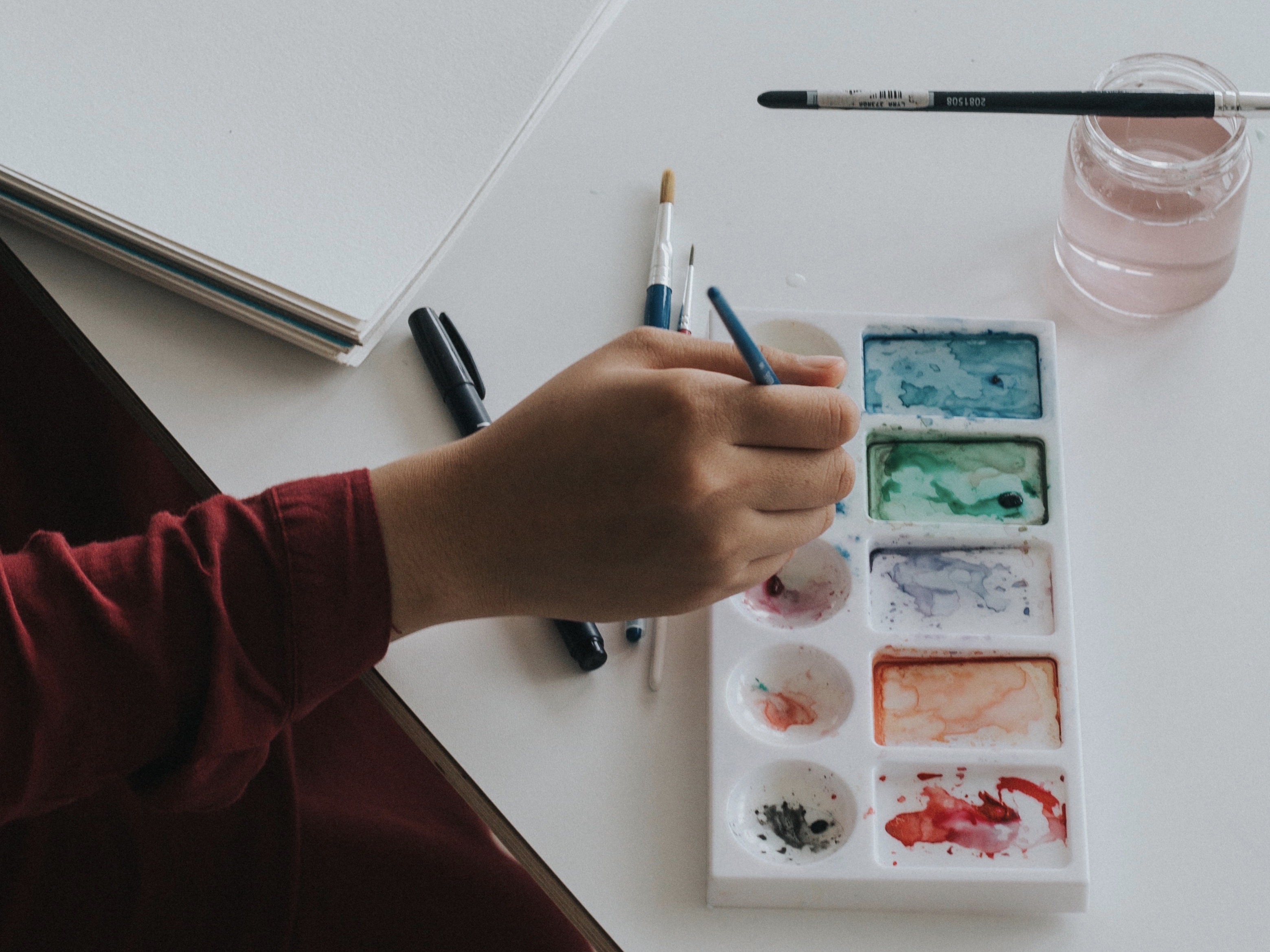 A watercolor paint palette with a hand holding a paintbrush poised over it.