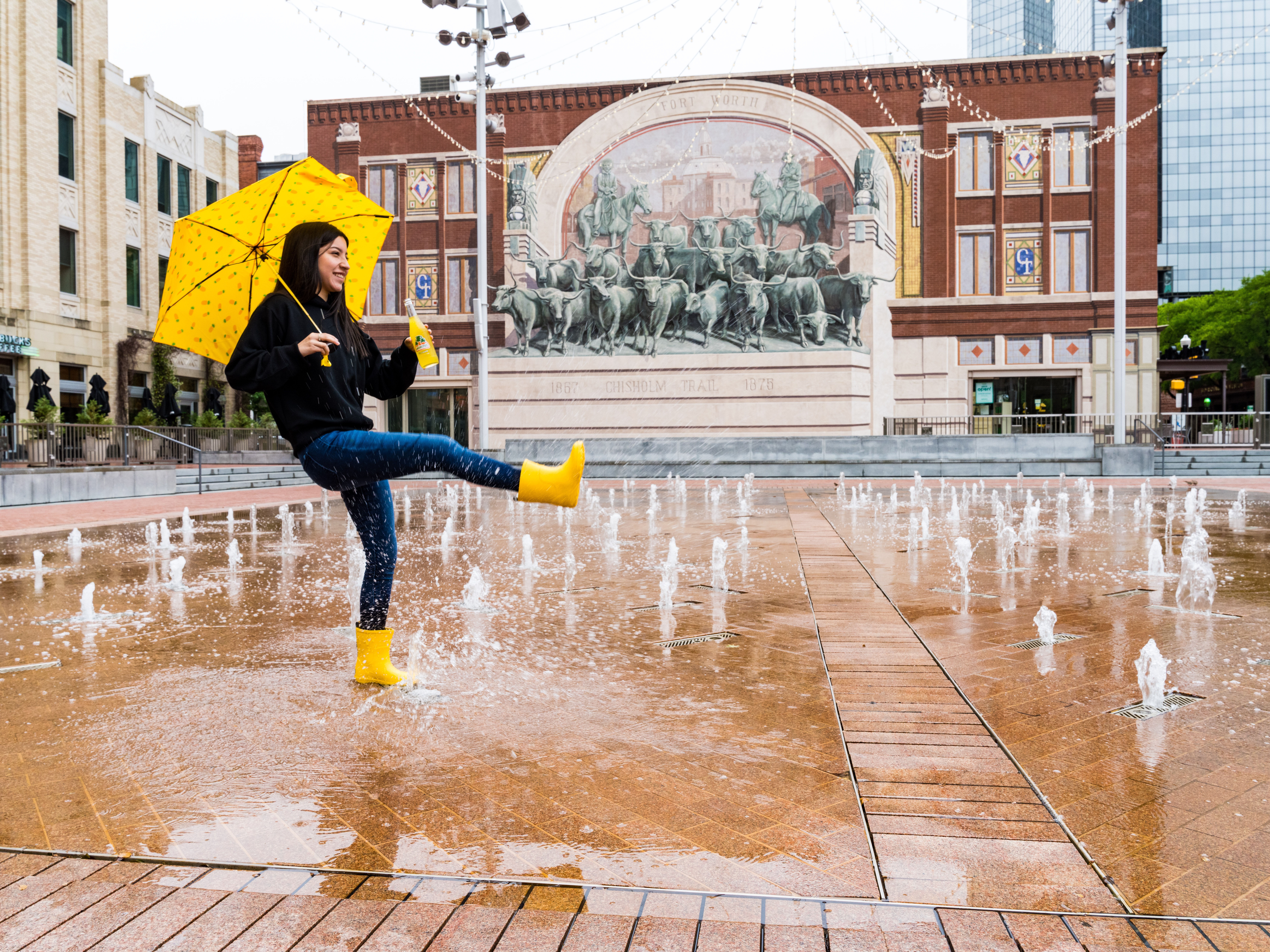 A young woman in yellow rainboots, with a yellow umbrella and bottle of soda, in a rainy plaza kicking one foot up while smiling.